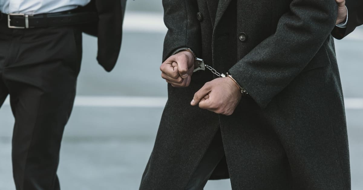 This photo depicts an individual wearing a dark suit and shackled with hand cuffs.